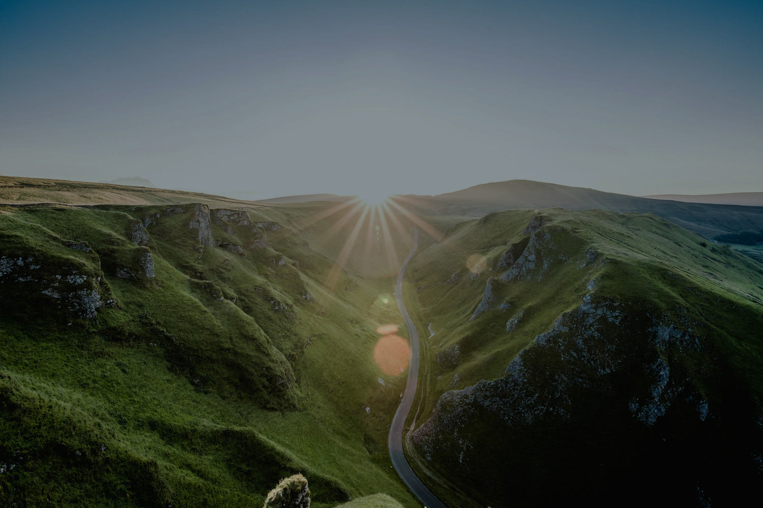 Sun starting to rise over green hills with a small paved road in between
