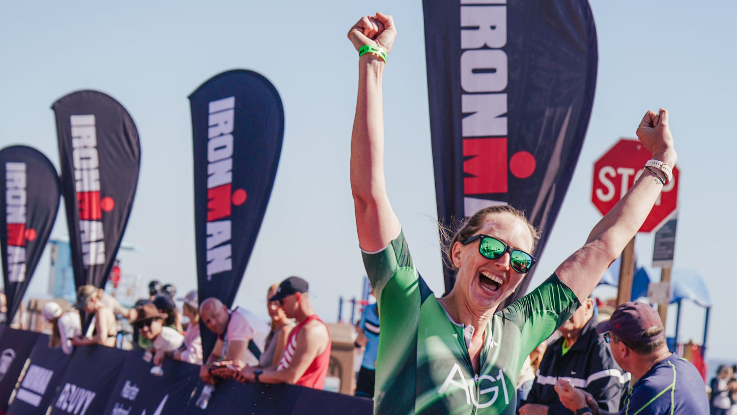 Person celebrating at an Ironman event with arms raised, wearing a green shirt and black shorts.